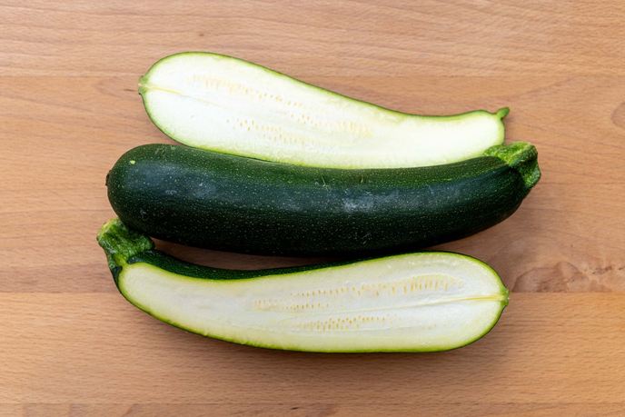 Drei Zucchini auf einem Holzbrett, zwei der Länge nach halbiert mit sichtbarem Kerngehäuse in der Mitte.